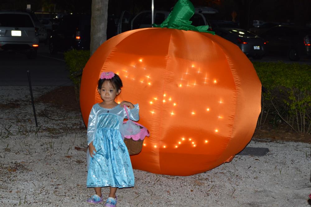 Young girl dressed in blue princess outfit with pink bow in hair and holding candy basket stands in front of large, inflatable, glowing jack-o-lantern